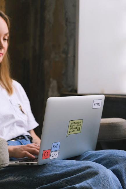 A person sitting outdoors on a patio or garden area, using a silver laptop with visible programming stickers including HTML, PHP, and a QR code. The individual is wearing a white shirt and blue jeans,
